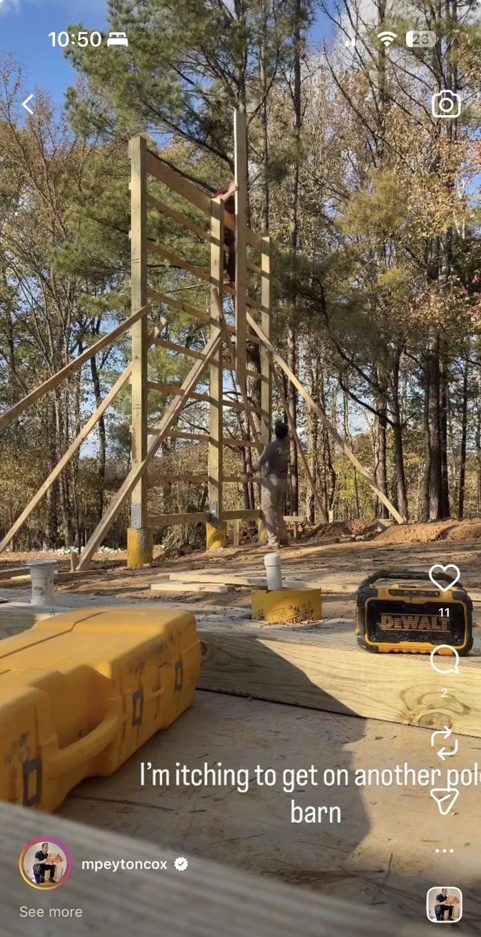 A timber frame structure under construction in a wooded lot, with fresh lumber stacked in the foreground.